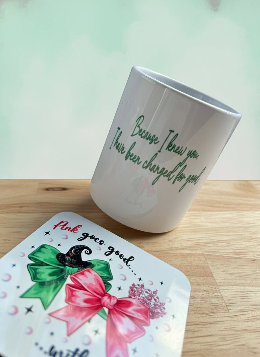 White mug with green text on a wooden surface next to a colorful coaster.
